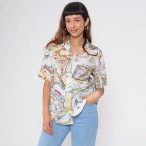 May include: A short-sleeved button-up shirt with a map print in shades of blue, yellow, and red. The shirt has a classic collar and is worn with light blue jeans. The map design features continents and nautical elements.
