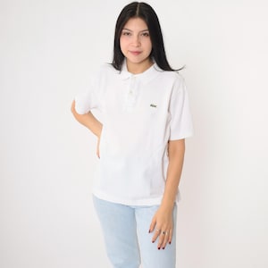 May include: A white polo shirt with short sleeves and a collar. The shirt has a small green crocodile logo on the left chest. The model is wearing light blue jeans. The background is plain white.