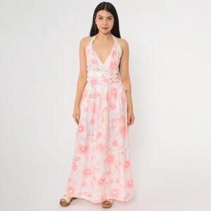 May include: A white halter-neck maxi dress with a pink floral pattern. The dress features a V-neckline, fitted waist, and a flowing skirt. The model is wearing the dress, standing in front of a white background.