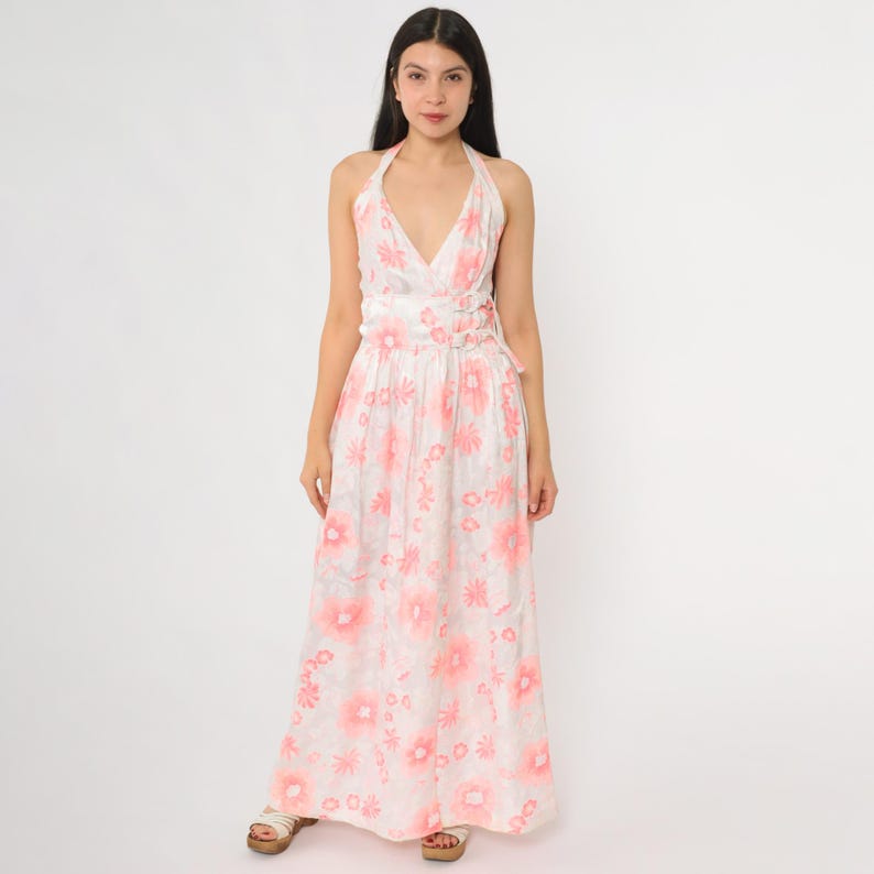 May include: A white halter-neck maxi dress with a pink floral pattern. The dress features a V-neckline, fitted waist, and a flowing skirt. The model is wearing the dress, standing in front of a white background.