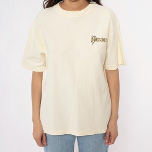 May include: Cream-colored t-shirt with short sleeves. The shirt features the text "GIRLSCOUT" in a stylized font on the upper right chest area. The shirt is made of a soft material and is worn by a person wearing blue jeans.