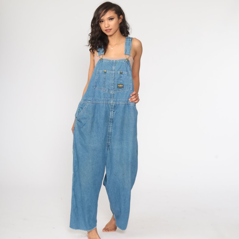 May include: A light blue denim overall dress with a wide leg and a relaxed fit. The straps are adjustable and have metal buckles. The overall dress has a front pocket and a back pocket.