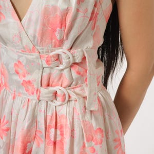 May include: A white dress with a pink floral pattern. The dress features a V-neck, a cinched waist with white buckle details, and a flowing skirt. The fabric has a subtle sheen.