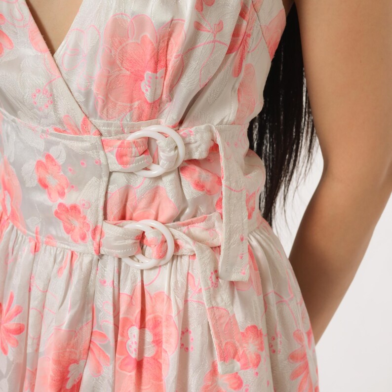 May include: A white dress with a pink floral pattern. The dress features a V-neck, a cinched waist with white buckle details, and a flowing skirt. The fabric has a subtle sheen.