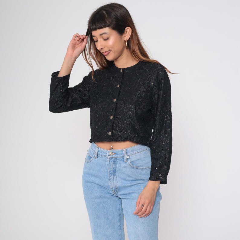 Puede incluir: Una camisa negra de manga larga con botones y un patr&oacute;n texturizado. La camisa tiene cuello redondo y est&aacute; recortada a la cintura. Los botones son de color marr&oacute;n claro. La persona de la imagen lleva vaqueros azules claros.