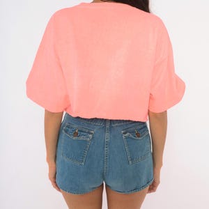 K&ouml;nnte beinhalten: Ein leuchtend neon-korallenfarbenes T-Shirt mit kurzen &Auml;rmeln, kombiniert mit mittelblauen Jeansshorts. Die Shorts verf&uuml;gen &uuml;ber zwei Ges&auml;&szlig;taschen mit gekn&ouml;pften Klappen und einen klassischen Saum, was einen l&auml;ssigen Look mit einem Oberteil ergibt.