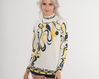 May include: A long-sleeve mock neck top in an off-white color featuring a bold, abstract graphic pattern in navy blue and bright yellow. The design includes flowing, organic shapes across the front and sleeves, with a structured border at the hem.
