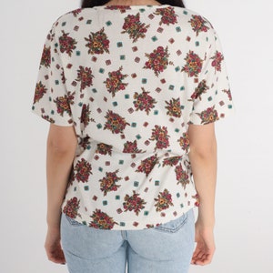 May include: A white short-sleeved shirt with a floral print. The flowers are in shades of red, pink, yellow, and green. The shirt has a scoop neck and a relaxed fit.