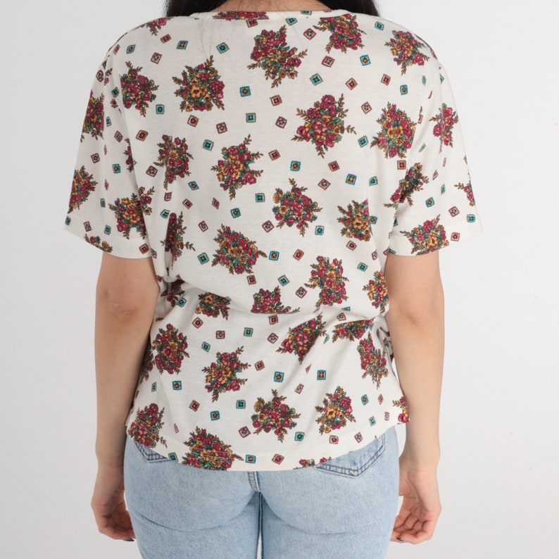 May include: A white short-sleeved shirt with a floral print. The flowers are in shades of red, pink, yellow, and green. The shirt has a scoop neck and a relaxed fit.