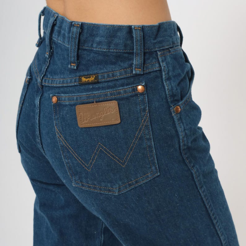 May include: Dark blue denim jeans with a Wrangler label on the back pocket. The jeans have copper-colored rivets and a classic five-pocket design. The Wrangler logo is visible on the back pocket and above the back pocket.