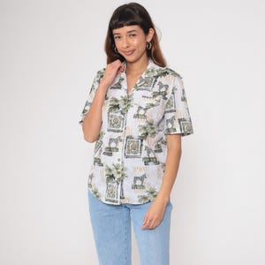 May include: A short-sleeved button-down shirt with a white background and a repeating pattern of zebras, palm trees, and geometric shapes in shades of green and black. The shirt is paired with light blue jeans.