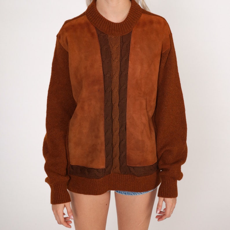 May include: A brown sweater with a suede panel on the front. The sweater has a cable knit design and a crew neck.