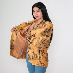 May include: A tan blazer with a deer and landscape pattern. The blazer has a peach-colored lining and long sleeves. The image shows the blazer open, revealing the lining. The blazer is worn with blue jeans.