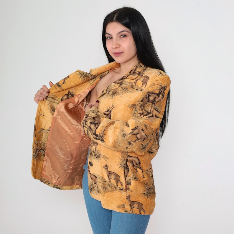May include: A tan blazer with a deer and landscape pattern. The blazer has a peach-colored lining and long sleeves. The image shows the blazer open, revealing the lining. The blazer is worn with blue jeans.