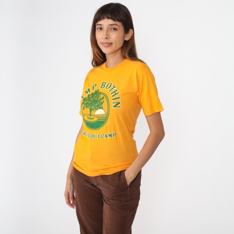 May include: Mustard yellow t-shirt with a green and white graphic that reads "CAMP BOTHIN RECOUT CAMP". The graphic features a tree, a setting sun, and a circular design. The shirt is short-sleeved and has a crew neck.