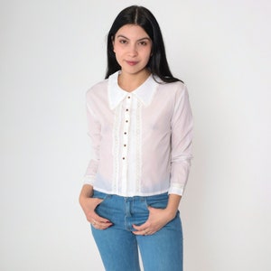 May include: A white long-sleeved button-up shirt with a lace trim and a large collar. The shirt is semi-sheer and has a cropped fit.