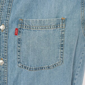 K&ouml;nnte beinhalten: Hellblaues Jeanshemd mit Knopfleiste und Brusttasche. Das Hemd hat silberne Kn&ouml;pfe und ein kleines rotes Etikett auf der Tasche. Der Stoff hat einen klassischen, abgenutzten Look.