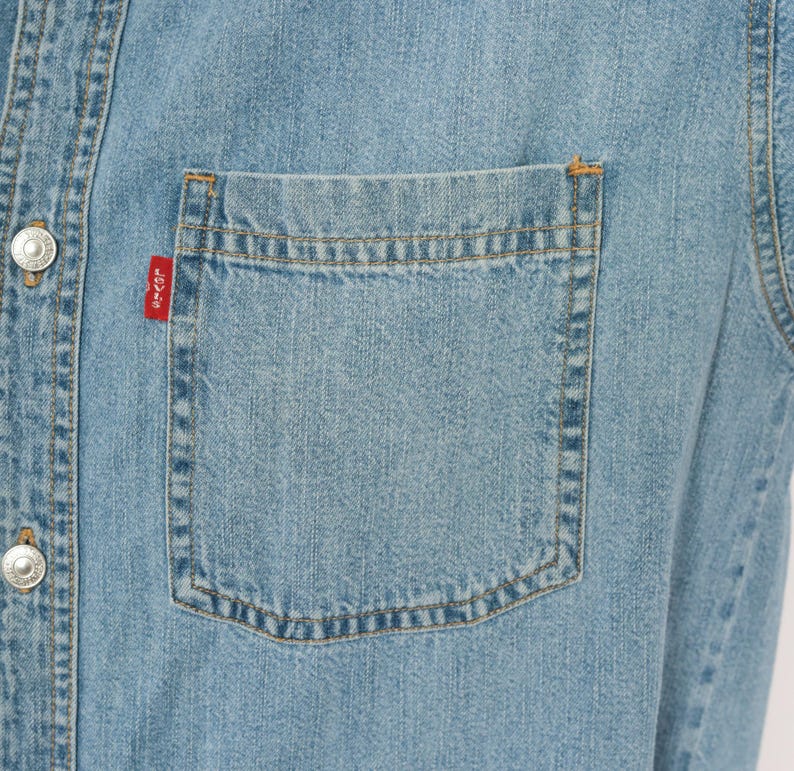 K&ouml;nnte beinhalten: Hellblaues Jeanshemd mit Knopfleiste und Brusttasche. Das Hemd hat silberne Kn&ouml;pfe und ein kleines rotes Etikett auf der Tasche. Der Stoff hat einen klassischen, abgenutzten Look.
