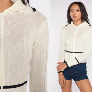 May include: Cream-colored zip-up sweater with a cable knit design. The sweater features navy blue trim on the collar, cuffs, and waist. The sweater has a collared neckline and long sleeves.