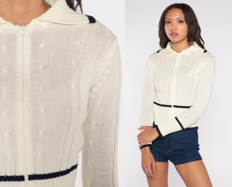 May include: Cream-colored zip-up sweater with a cable knit design. The sweater features navy blue trim on the collar, cuffs, and waist. The sweater has a collared neckline and long sleeves.