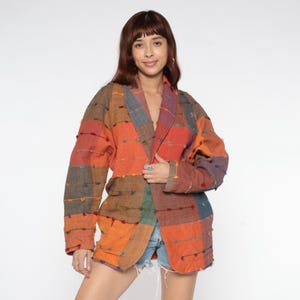 May include: A patchwork blazer in shades of orange, red, brown, and blue. The jacket has a relaxed fit and features a shawl collar. The fabric appears to be textured, with small, colorful embellishments. The blazer is open, revealing the wearer's outfit.