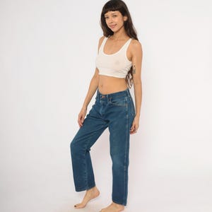 May include: A woman wearing a white crop top and blue jeans. The jeans are a classic style with a straight leg and a medium wash. The top is a simple tank style.