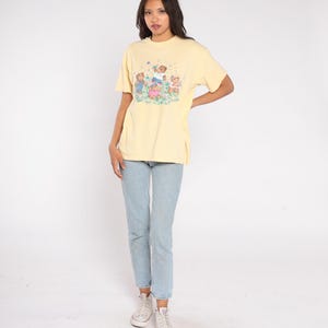 May include: A light yellow t-shirt featuring a graphic of children playing in a field. The shirt is paired with light blue jeans and white sneakers. The t-shirt has short sleeves and a crew neck.