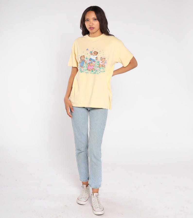 May include: A light yellow t-shirt featuring a graphic of children playing in a field. The shirt is paired with light blue jeans and white sneakers. The t-shirt has short sleeves and a crew neck.
