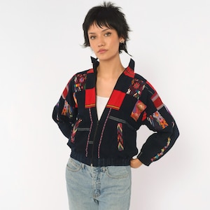 May include: A black zip-up jacket featuring a V-neckline and long sleeves. The garment is decorated with various rectangular patches in red and multi-colored woven patterns. The jacket has a ribbed hem and cuffs, and is worn over a white shirt.