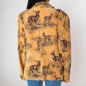 May include: A tan blazer with a repeating pattern of deer in a landscape scene. The blazer has a classic collar and a slightly textured fabric. The deer are depicted in shades of brown and black against a tan background.