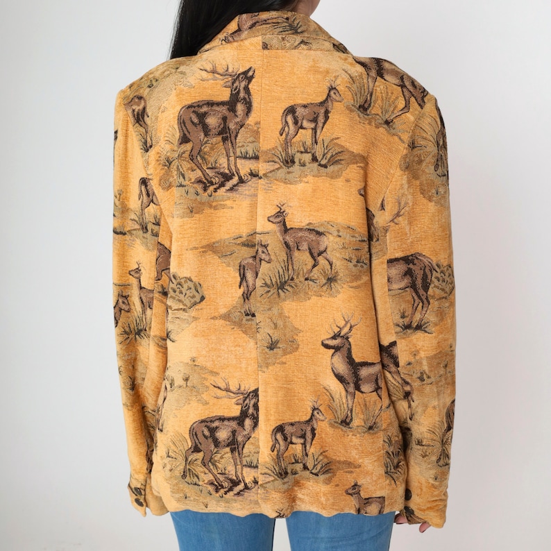 May include: A tan blazer with a repeating pattern of deer in a landscape scene. The blazer has a classic collar and a slightly textured fabric. The deer are depicted in shades of brown and black against a tan background.