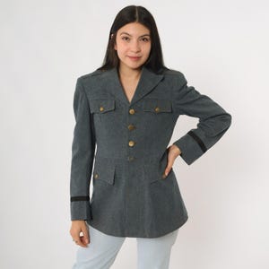 K&ouml;nnte beinhalten: Graue Jacke im Milit&auml;rstil mit goldenen Kn&ouml;pfen und Taschen. Die Jacke hat eine taillierte Passform, lange &Auml;rmel mit schwarzen Streifen an den Manschetten und Schulterklappen. Ein Vintage-inspiriertes Kleidungsst&uuml;ck.