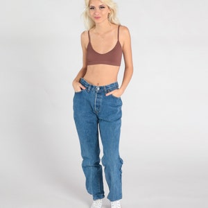 May include: A woman wearing a brown ribbed bralette and blue denim jeans with a high waist. She is wearing white trainers.