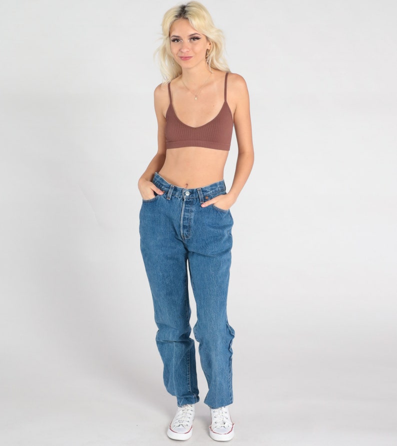 May include: A woman wearing a brown ribbed bralette and blue denim jeans with a high waist. She is wearing white trainers.