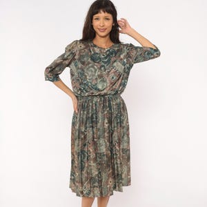 May include: A midi-length dress featuring a round neckline, three-quarter length sleeves, and an elasticized waist. The garment is patterned with an abstract, multi-colored print in shades of muted green, brown, and grey with a pleated skirt.