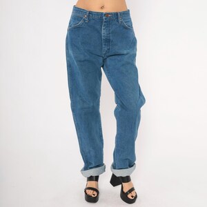 May include: Blue denim jeans with a button and zipper closure, featuring a classic five-pocket design. The jeans have a relaxed fit and are cuffed at the ankles. The model is wearing black platform sandals.