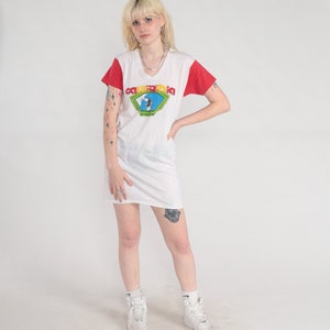 K&ouml;nnte beinhalten: Wei&szlig;es T-Shirt-Kleid mit roten &Auml;rmeln und einem Grafikdruck eines Surfers mit dem Text "California" in Gelb, Rot und Blau.