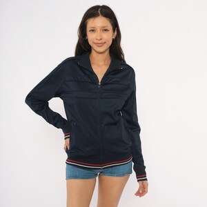 May include: A dark navy blue zip-up track jacket featuring a high collar, long sleeves with ribbed cuffs, and horizontal stitched detailing across the chest. The hem and cuffs are accented with thin white and red stripes, and the front has a zipper.
