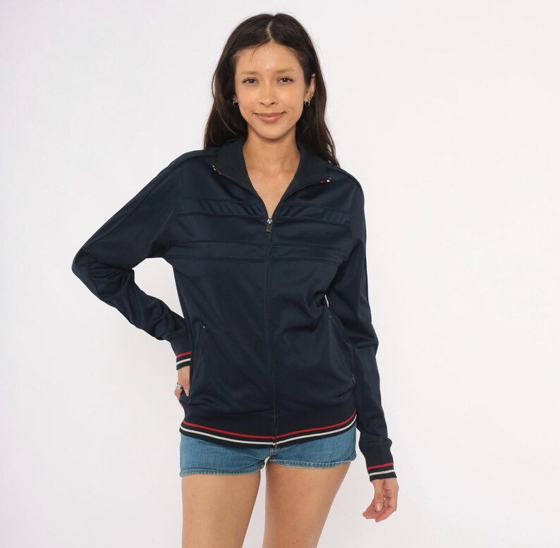 May include: A dark navy blue zip-up track jacket featuring a high collar, long sleeves with ribbed cuffs, and horizontal stitched detailing across the chest. The hem and cuffs are accented with thin white and red stripes, and the front has a zipper.