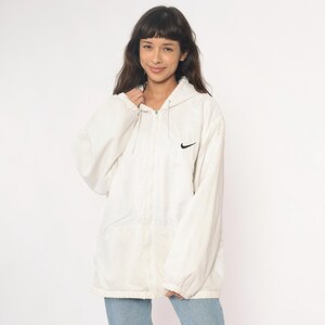 Puede incluir: Chaqueta con capucha Nike blanca con el logotipo negro de Nike en el pecho. La chaqueta tiene cierre de cremallera completa y capucha con cord&oacute;n. La chaqueta se lleva sobre unos vaqueros azules.