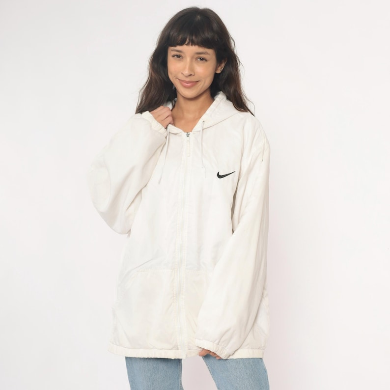 Puede incluir: Chaqueta con capucha Nike blanca con el logotipo negro de Nike en el pecho. La chaqueta tiene cierre de cremallera completa y capucha con cord&oacute;n. La chaqueta se lleva sobre unos vaqueros azules.