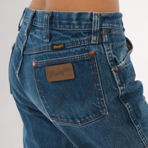 Pode incluir: Close-up de jeans de ganga azul com o log&oacute;tipo Wrangler no bolso traseiro. Os jeans t&ecirc;m rebites cor de cobre e um design cl&aacute;ssico de cinco bolsos. A ganga tem um aspeto desbotado.