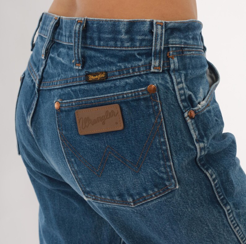 Pode incluir: Close-up de jeans de ganga azul com o log&oacute;tipo Wrangler no bolso traseiro. Os jeans t&ecirc;m rebites cor de cobre e um design cl&aacute;ssico de cinco bolsos. A ganga tem um aspeto desbotado.