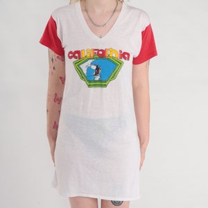 K&ouml;nnte beinhalten: Wei&szlig;es Langarm-T-Shirt mit roten &Auml;rmeln und einem Grafikdruck eines Surfers in einer blauen und gr&uuml;nen Welle mit dem Wort "California" in einer Regenbogen-Schrift.