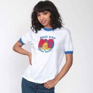 Könnte beinhalten: Weißes Ringer-T-Shirt mit blauem Besatz und dem Text "Just Say NO" in Rot und Blau, mit einer Abbildung des Kapitols. Das Shirt ist im Vintage-Stil.