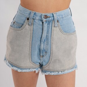Puede incluir: Pantalones cortos de mezclilla azul claro con un panel de mezclilla gris contrastante a cada lado. Los pantalones cortos tienen cintura alta y dobladillo deshilachado.