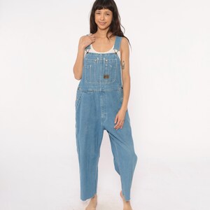 May include: Light blue denim overalls with adjustable straps, multiple pockets, and a brown leather patch. The overalls have a relaxed fit and are made of a durable denim material. A white tank top is worn underneath.