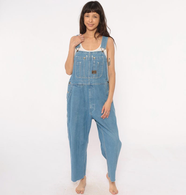 May include: Light blue denim overalls with adjustable straps, multiple pockets, and a brown leather patch. The overalls have a relaxed fit and are made of a durable denim material. A white tank top is worn underneath.