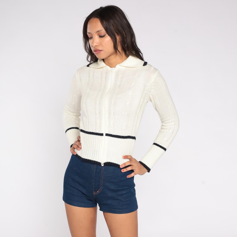 May include: Cream-colored zip-up sweater with black trim on the collar, cuffs, and waist. The sweater has a cable knit design and is paired with dark blue denim shorts. The outfit is a vintage style.