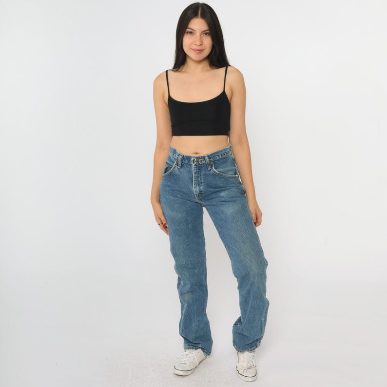 Puede incluir: Una persona con un top corto negro con tirantes finos y vaqueros azules. Los vaqueros tienen un dise&ntilde;o cl&aacute;sico de cinco bolsillos y un corte relajado. Zapatillas blancas completan el conjunto.
